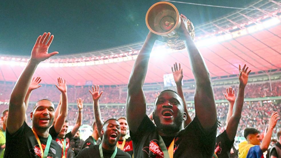 Leverkusens Victor Boniface (r) hält den Pokal jubelnd hoch, während seine Teamkollegen feiern. Foto: Federico Gambarini/dpa