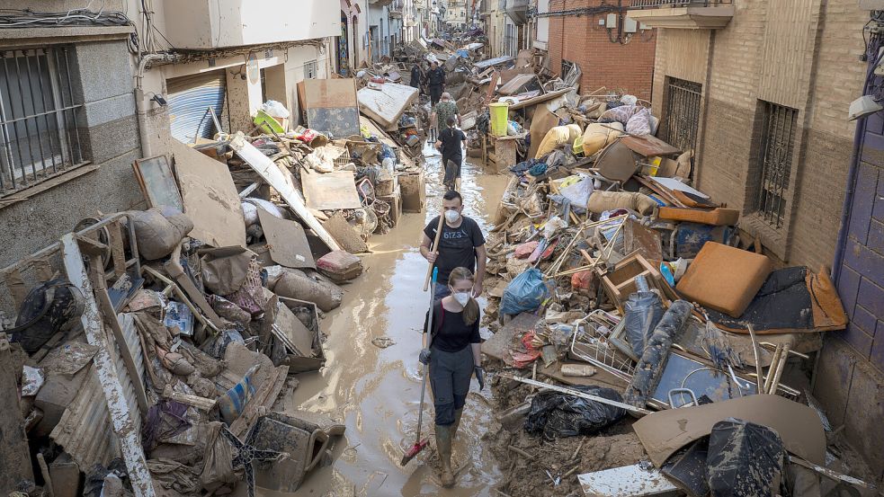 Menschen gehen durch eine Straße mit aufgetürmten Möbeln und Müll an den Seiten in einem von den Überschwemmungen vor einer Woche betroffenen Gebiet in Valencia. Noch immer gelten zahlreiche Personen als vermisst. Foto: dpa/Emilio Morenatti