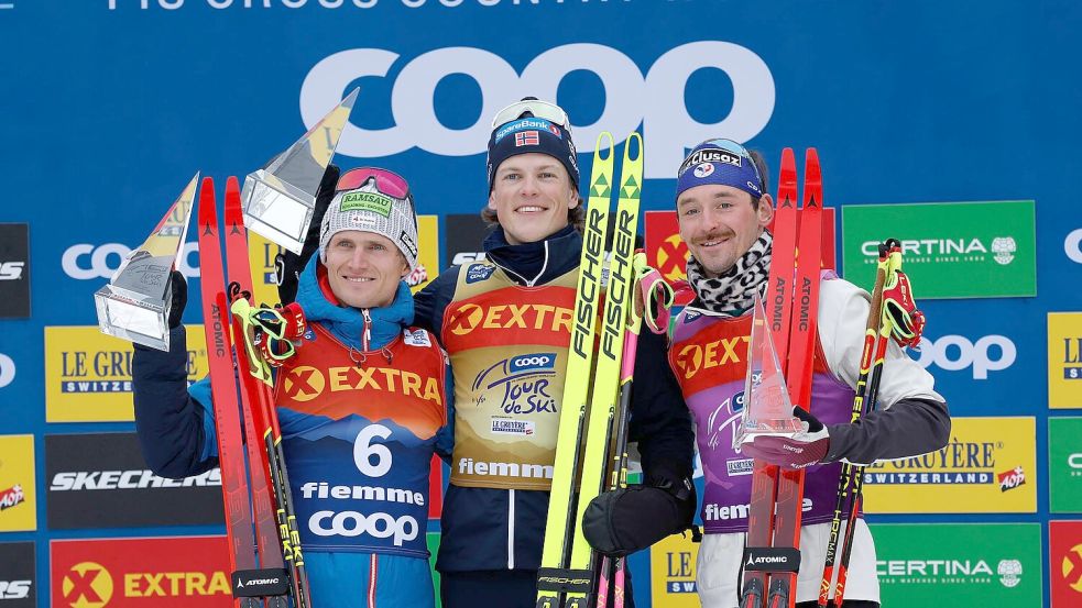 Johannes Hoesflot Klaebo (M) aus Norwegen jubelt neben dem Zweitplatzierten Mika Vermeulen (l) aus Österreich und dem Drittplatzierten Hugo Lapalus aus Frankreich auf dem Podium der Tour de Ski. Foto: Alessandro Trovati/AP/dpa